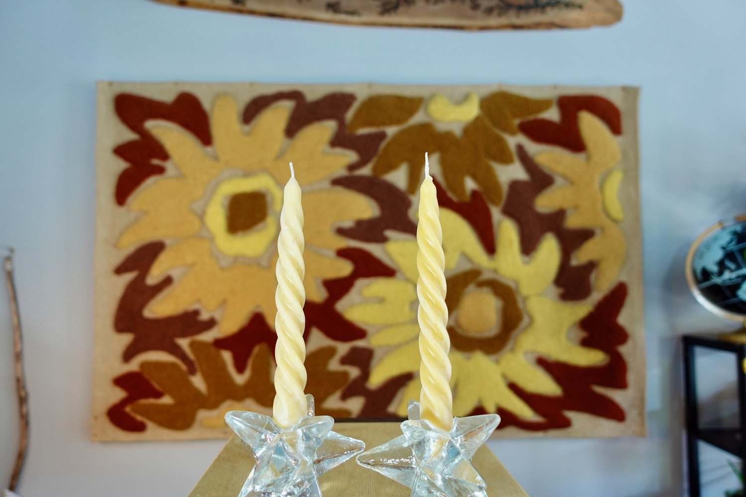 Two beeswax tapers in glass holders in front of a sunflower hooked rug
